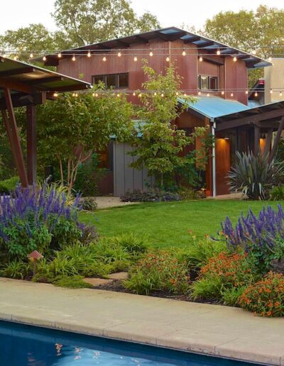 A well-manicured backyard with a swimming pool, string lights, and a variety of plants.