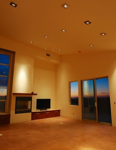 Modern living room at dusk with large windows and recessed ceiling lighting.
